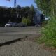 Low-angle street scene with cars driving on a paved road and tall green grass growing along the right edge, city buildings in the background.