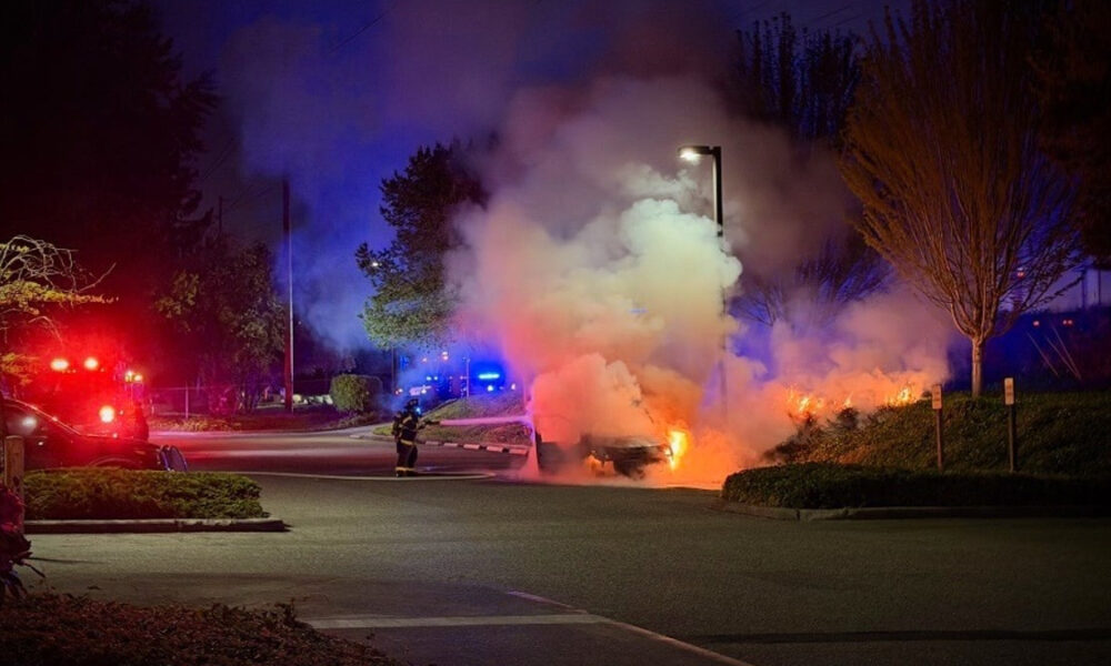 Car on fire in a parking lot at night, firefighters spraying flames amid red and blue emergency lights.