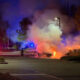 Car on fire in a parking lot at night, firefighters spraying flames amid red and blue emergency lights.