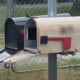 Beige mailbox with a red flag on a metal post, its curved lid open and the inner slot visible near a chain-link fence.