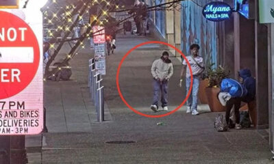 Man in light gray hoodie and dark pants stands with hands in pockets on a city sidewalk, circled in red. Two other people nearby.