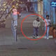 Man in light gray hoodie and dark pants stands with hands in pockets on a city sidewalk, circled in red. Two other people nearby.