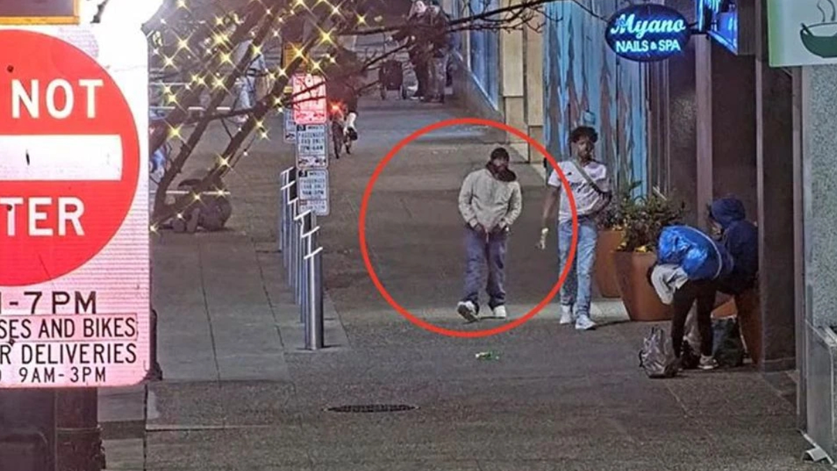 Man in light gray hoodie and dark pants stands with hands in pockets on a city sidewalk, circled in red. Two other people nearby.