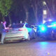 Nighttime street scene with a gray sedan stopped on the curb, police car flashing blue and red lights, and a group of people talking near the trunk.