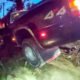 Dark SUV tilted in a ditch with 44 on its side, rear lights on, mud and vegetation around at dusk/dawn.