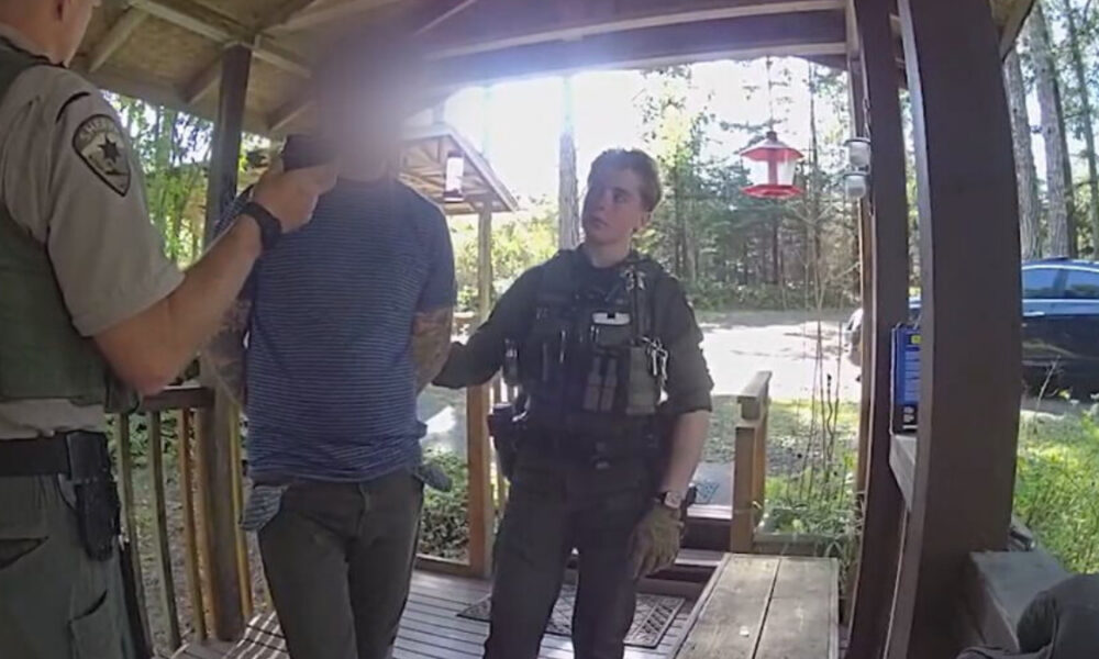 Two uniformed officers with tactical vests detain a man in a striped shirt on a wooden porch.