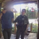 Two uniformed officers with tactical vests detain a man in a striped shirt on a wooden porch.
