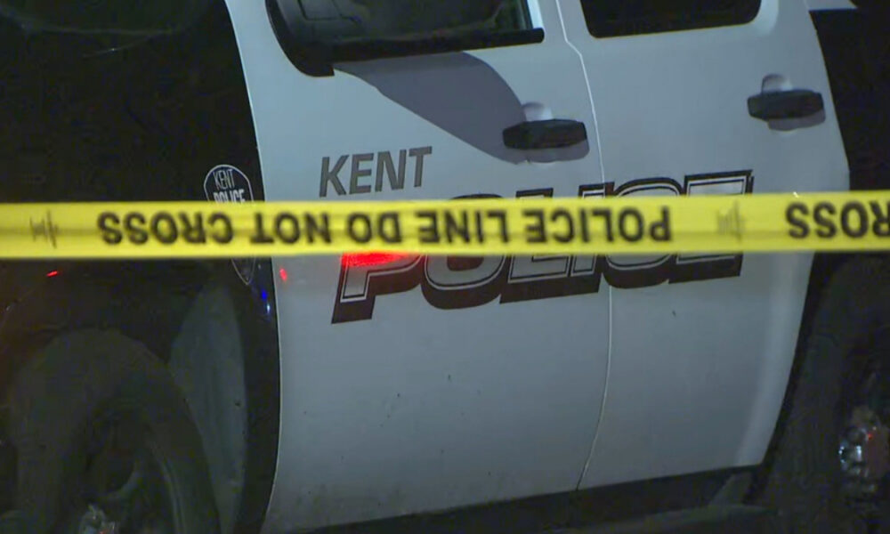 Nighttime police car with 'POLICE' lettering and 'KENT' visible behind yellow 'POLICE LINE DO NOT CROSS' tape. Crime scene context.