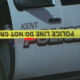 Nighttime police car with 'POLICE' lettering and 'KENT' visible behind yellow 'POLICE LINE DO NOT CROSS' tape. Crime scene context.