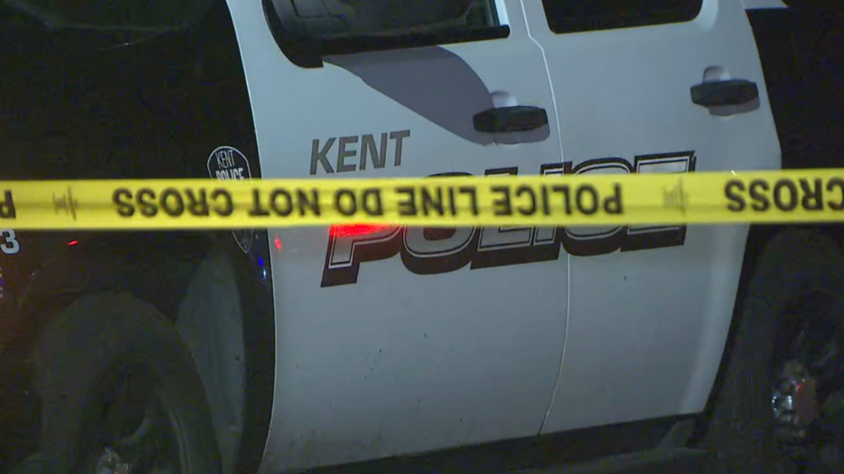 Nighttime police car with 'POLICE' lettering and 'KENT' visible behind yellow 'POLICE LINE DO NOT CROSS' tape. Crime scene context.