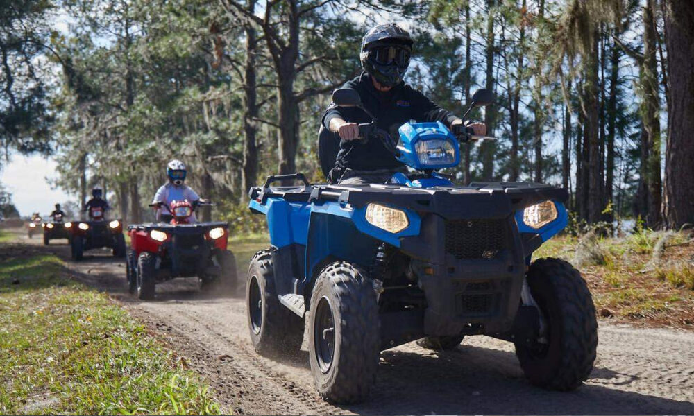 Group of people riding ATVs on a dusty forest trail, with a blue ATV in the foreground and trees on both sides.