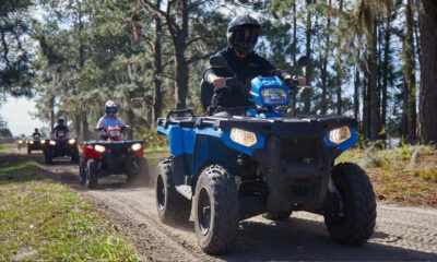 Group of people riding ATVs on a dusty forest trail, with a blue ATV in the foreground and trees on both sides.