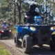Group of people riding ATVs on a dusty forest trail, with a blue ATV in the foreground and trees on both sides.