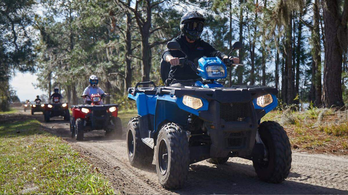 Group of people riding ATVs on a dusty forest trail, with a blue ATV in the foreground and trees on both sides.