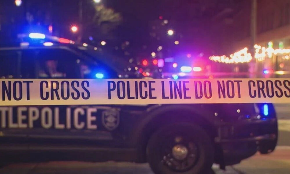 Nighttime police car behind yellow 'Do Not Cross' tape marking a crime scene with flashing lights in the background, streets dimly lit by streetlights and decorations.