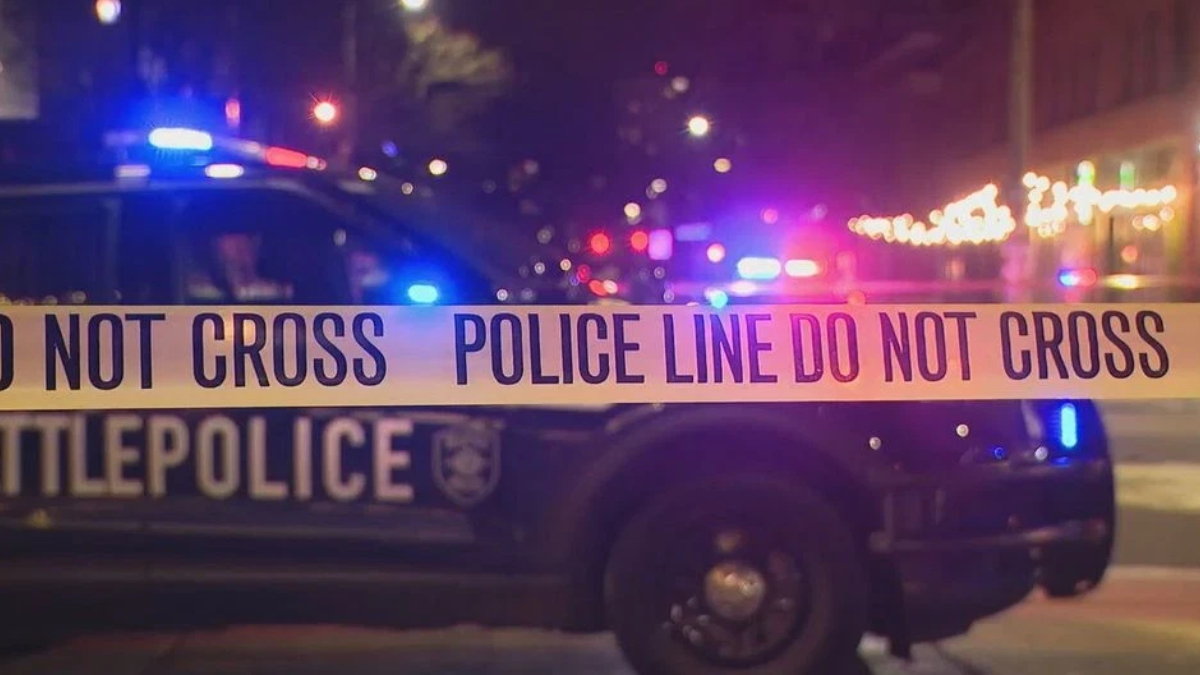 Nighttime police car behind yellow 'Do Not Cross' tape marking a crime scene with flashing lights in the background, streets dimly lit by streetlights and decorations.