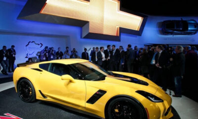 Yellow Chevrolet Corvette on display at an auto show with a large Chevrolet logo backdrop and a crowd of attendees behind it? and photographers nearby.