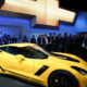 Yellow Chevrolet Corvette on display at an auto show with a large Chevrolet logo backdrop and a crowd of attendees behind it? and photographers nearby.