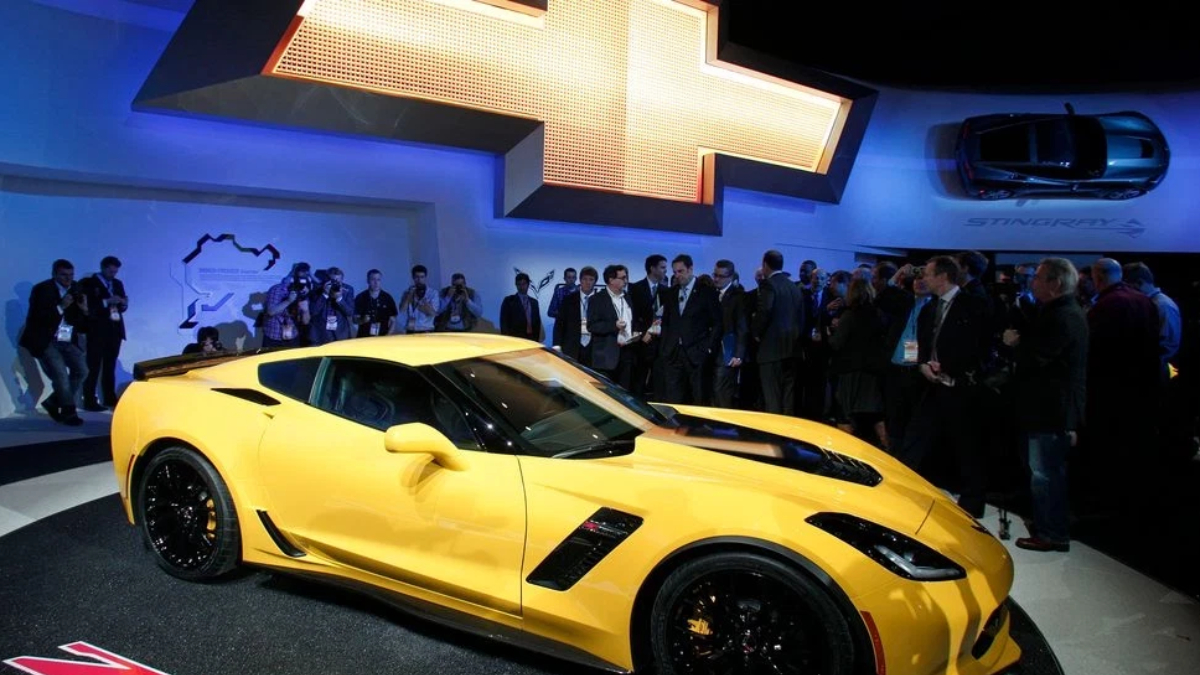 Yellow Chevrolet Corvette on display at an auto show with a large Chevrolet logo backdrop and a crowd of attendees behind it? and photographers nearby.