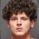 Mugshot of a young man with curly brown hair and neck tattoos, neutral expression