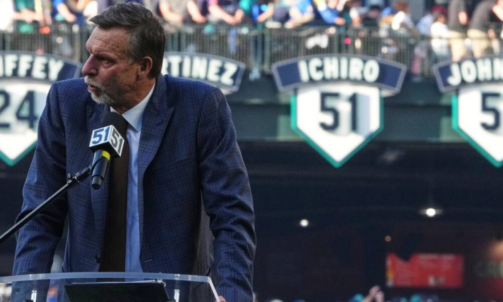 Man in a blue checkered suit speaks at a podium with a microphone, stadium banners and a crowd in the background.