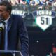 Man in a blue checkered suit speaks at a podium with a microphone, stadium banners and a crowd in the background.