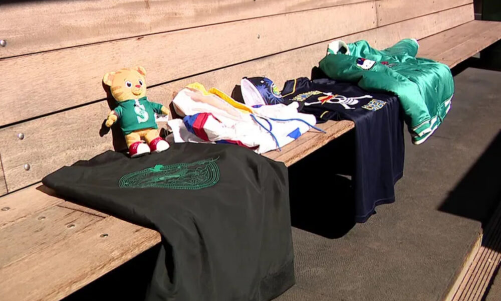 A stuffed bear in a green shirt sits on a wooden bench beside folded clothes and a dark T‑shirt with a green emblem.