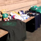 A stuffed bear in a green shirt sits on a wooden bench beside folded clothes and a dark T‑shirt with a green emblem.