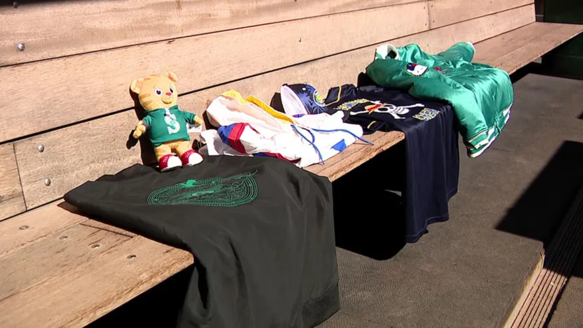 A stuffed bear in a green shirt sits on a wooden bench beside folded clothes and a dark T‑shirt with a green emblem.