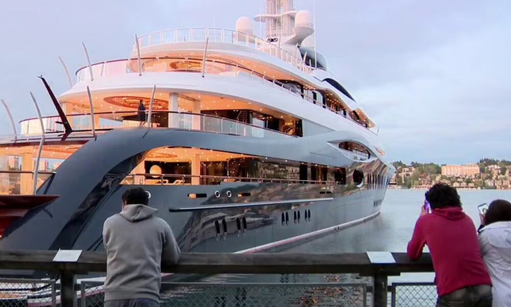A large luxury yacht docked at a marina with two people standing on a railing taking photos.