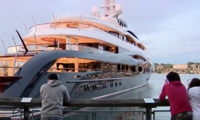 A large luxury yacht docked at a marina with two people standing on a railing taking photos.