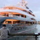 A large luxury yacht docked at a marina with two people standing on a railing taking photos.