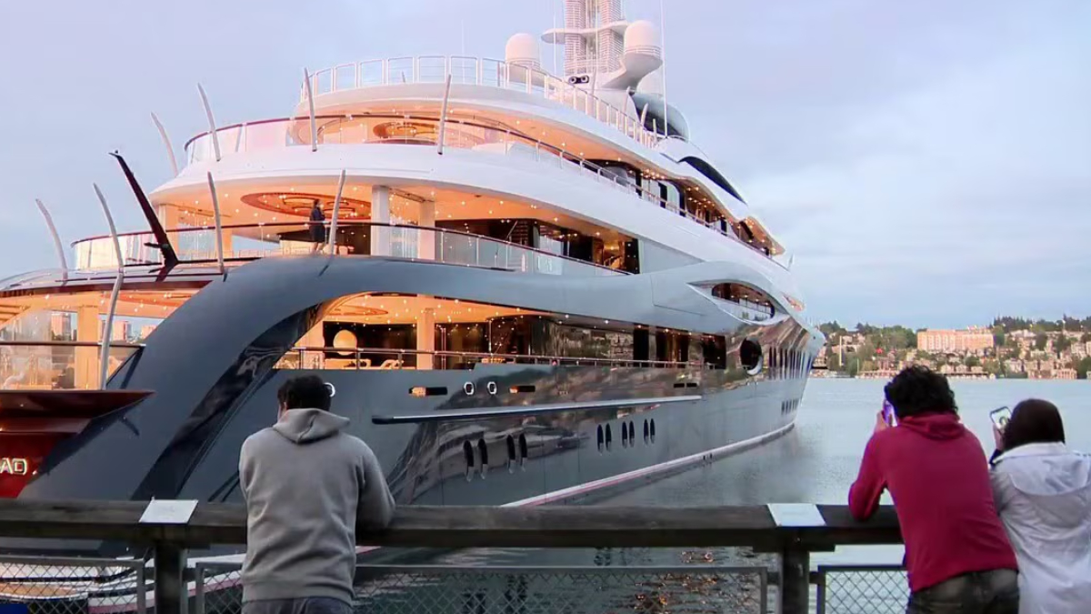 A large luxury yacht docked at a marina with two people standing on a railing taking photos.
