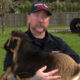 Firefighter in a dark uniform holding a brown and black goat in a grassy yard with a wooden fence behind him.