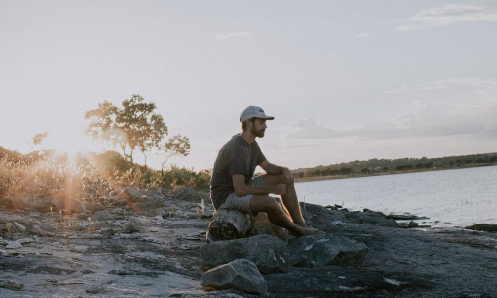 Man sits on rocks along a lakeshore at sunset, wearing a cap and gray shirt, gazing at the water.