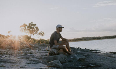 Man sits on rocks along a lakeshore at sunset, wearing a cap and gray shirt, gazing at the water.