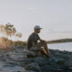 Man sits on rocks along a lakeshore at sunset, wearing a cap and gray shirt, gazing at the water.
