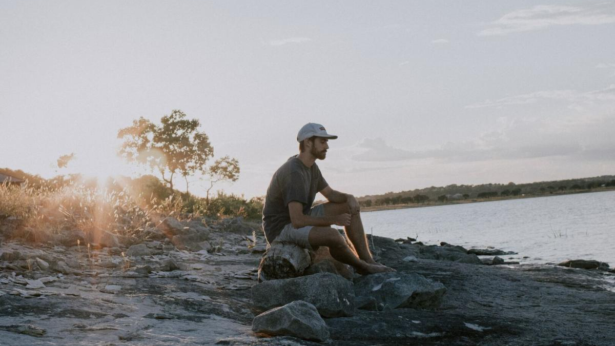 Man sits on rocks along a lakeshore at sunset, wearing a cap and gray shirt, gazing at the water.
