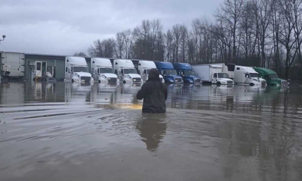 Person wading in floodwater in a truck yard with a row of semi-trailers parked along a building row in the distance.