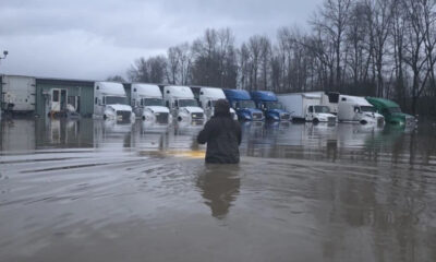 Person wading in floodwater in a truck yard with a row of semi-trailers parked along a building row in the distance.