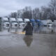 Person wading in floodwater in a truck yard with a row of semi-trailers parked along a building row in the distance.