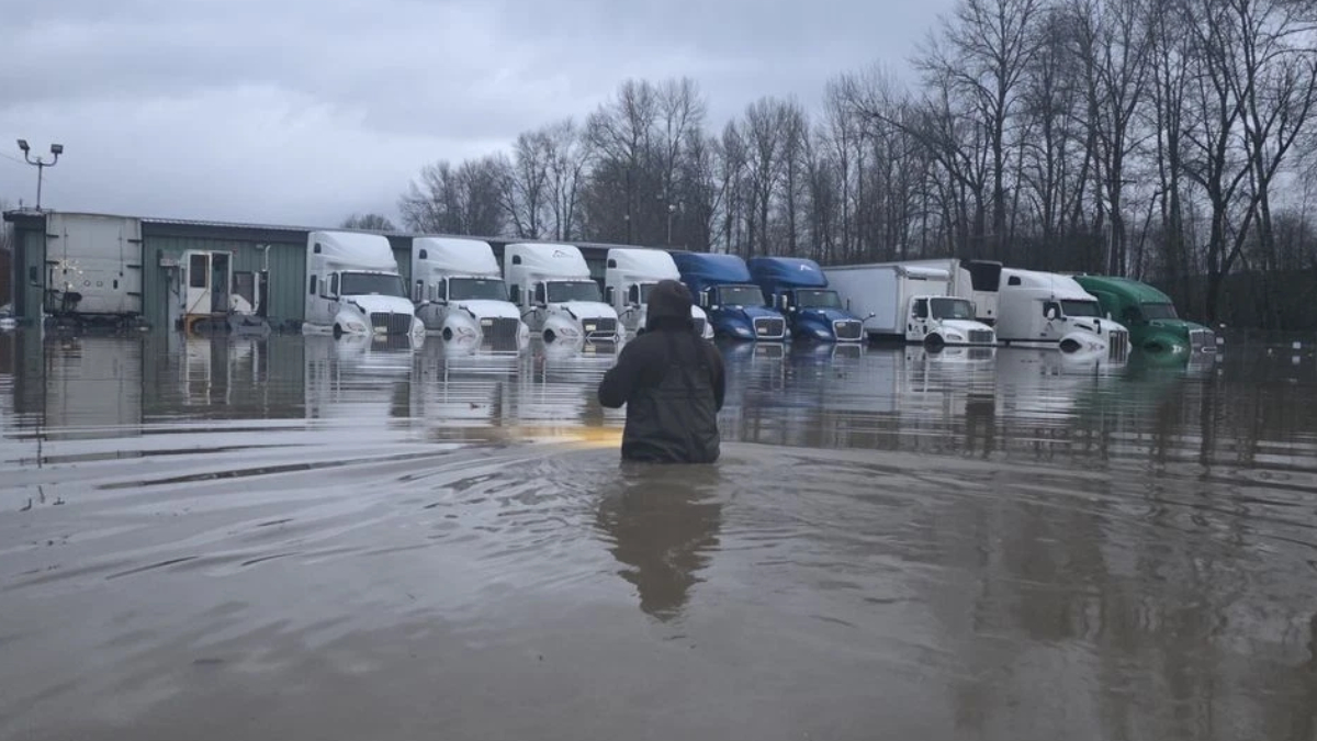 Person wading in floodwater in a truck yard with a row of semi-trailers parked along a building row in the distance.