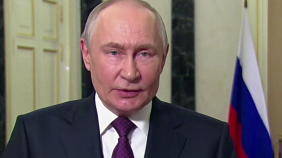 Close-up of a man in a dark suit speaking indoors, with a Russian flag in the background.