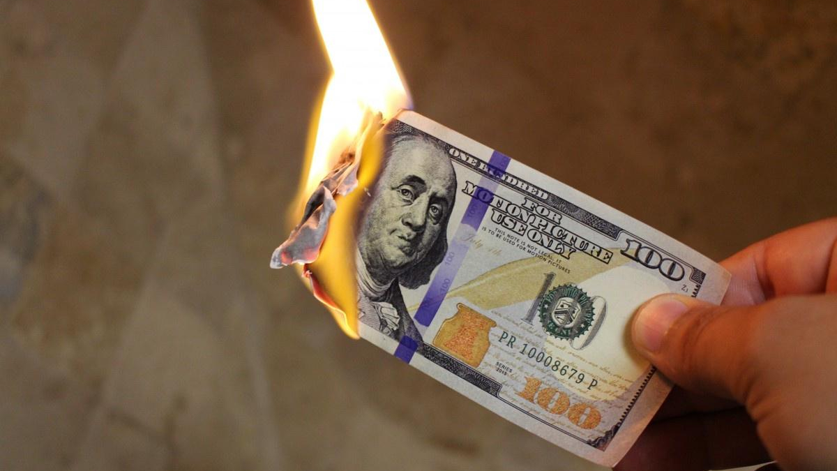 A hand holds a burning one-hundred-dollar bill with flames at the left side of the note of fire.