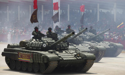Several armored tanks rolling in a public parade, with soldiers in the turrets and a smoky atmosphere as a crowded audience watches from behind barriers.