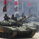 Several armored tanks rolling in a public parade, with soldiers in the turrets and a smoky atmosphere as a crowded audience watches from behind barriers.