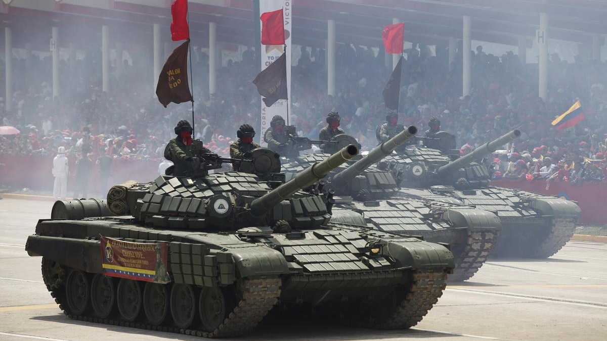 Several armored tanks rolling in a public parade, with soldiers in the turrets and a smoky atmosphere as a crowded audience watches from behind barriers.