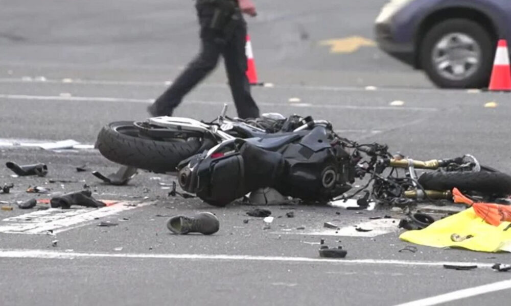 Motorcycle lying on its side in a road crash with scattered debris and a patrol car cone in the background.