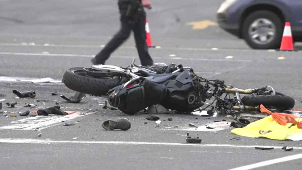 Motorcycle lying on its side in a road crash with scattered debris and a patrol car cone in the background.