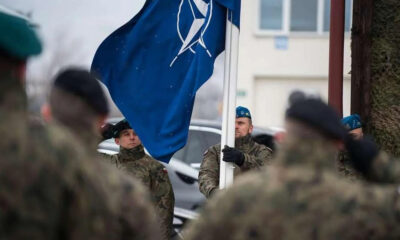 Soldier in camouflage holds a blue flag with a white emblem during a military ceremony, with colleagues in uniform nearby.
